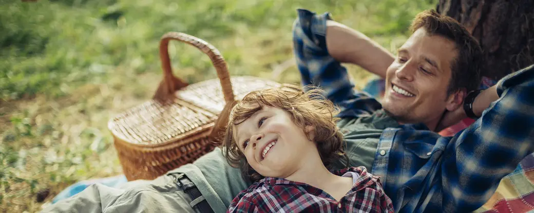 На изображении отец и сын лежат на траве и смеются, наслаждаясь видом друг друга. Оба одеты в клетчатые рубашки. Рядом с ними находится плетеная корзина. Атмосфера спокойная и радостная, передающая момент близости и семейного счастья.