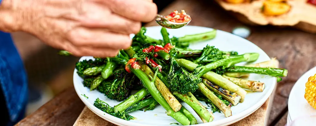 На тарелке лежат разные виды зеленых овощей, таких как брокколи и молодая спаржа, политыми соусом из перца. Видно, что кто-то подносит ложку с соусом, готовясь добавить его к блюду.
