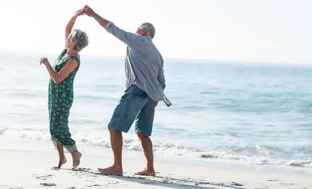 Два пожилых человека танцуют на берегу моря. Женщина в зеленом платье улыбается и держится за руки с мужчиной, который в светлой рубашке. Оба выглядят счастливыми, наслаждаясь моментом под солнечным небом и у моря.