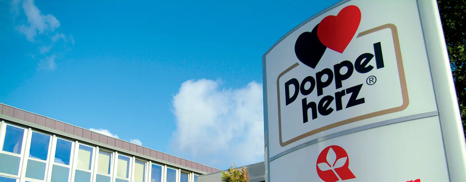 На изображении видно рекламное табло с логотипом компании "Doppelherz". Фон ясный, голубое небо с облаками. Логотип включает два сердца, одно красное, другое черное, и название компании, написанное крупными буквами.
