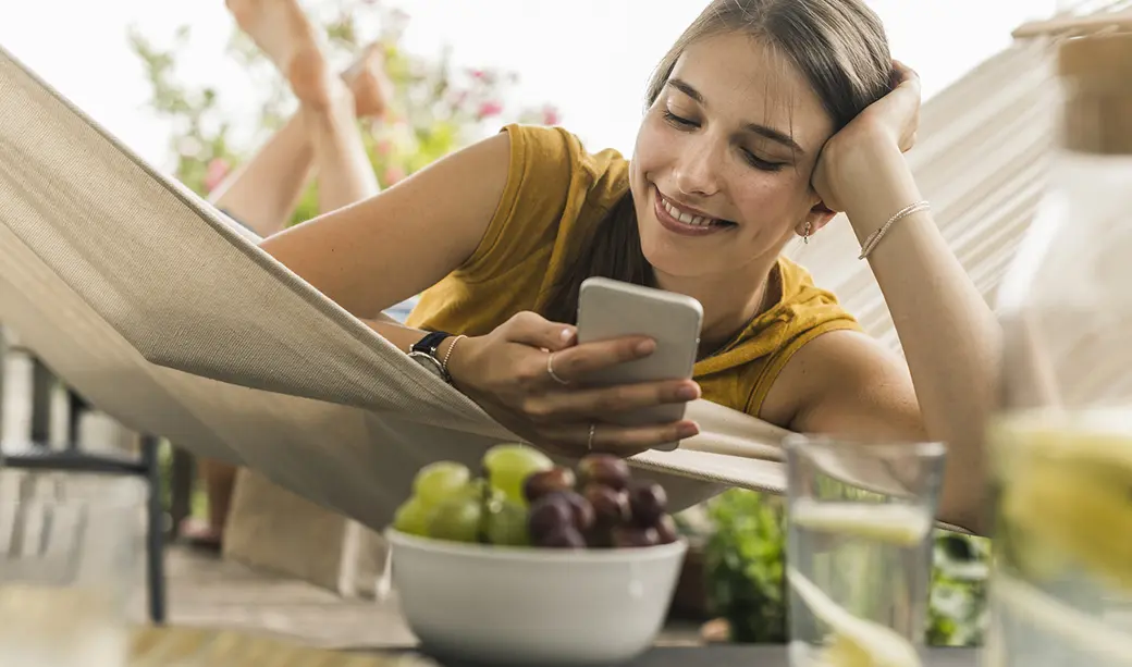 Молодая женщина лежит в гамаке и улыбается, смотря на телефон. Перед ней стоит миска с виноградом и зелеными ягодами. На заднем плане видны растения, создающие атмосферу уюта и спокойствия.