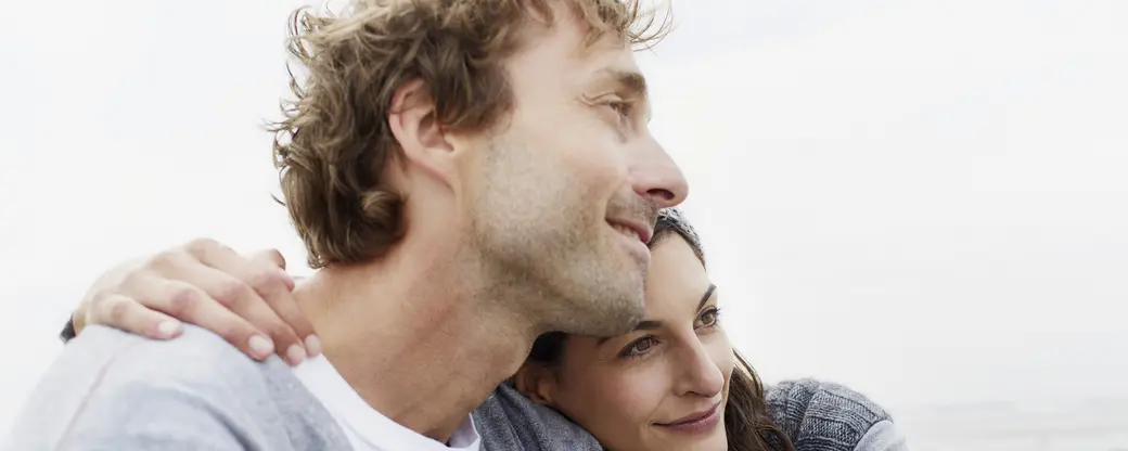 На этом изображении счастливая пара, уютно обнявшись, смотрит на море. Женщина нежно прижимается к мужчине, выражая тепло и близость. Фон слегка размытый с легкими облаками, создавая атмосферу спокойствия и гармонии.