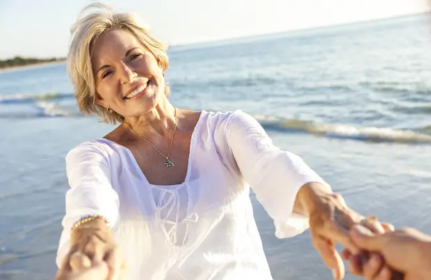 На фотографии женщина с короткими светлыми волосами улыбается, протягивая руки вперед. Она одета в белую блузу и стоит на берегу моря, на фоне спокойных волн и ясного неба. Атмосфера радости и дружелюбия.