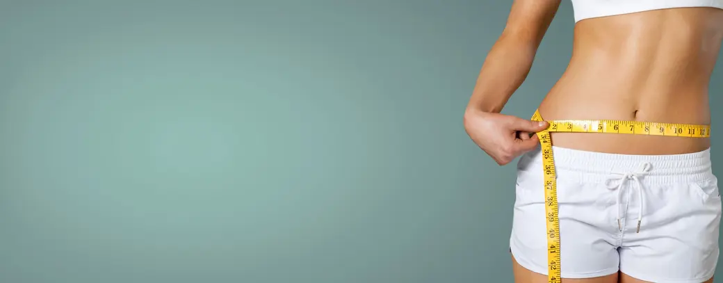 Женская фигура, показывающая талию, обернутую measuring tape. Вид сзади, женщина в белом топе и шортах. Изображение акцентирует внимание на измерении талии, что может символизировать фитнес или контроль веса. Фон светлый, нейтральный.