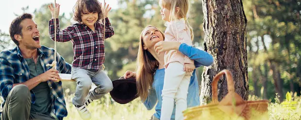 Счастливая семья на прогулке на природе. Мать держит на руках маленькую девочку, отец улыбается, а сын в клетчатой рубашке играет на качелях. Вокруг зелень деревьев, создающая атмосферу радости и единства.